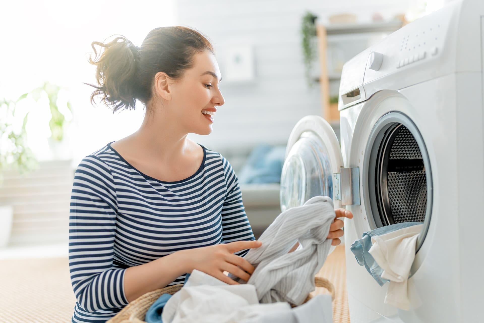 Woman loading clothes into a modern front-loading washing machine in a bright laundry room at home.